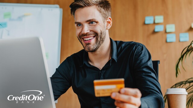 A young man smiles as he sits at his desk holding a credit card and using a laptop, which is a great tool for experiencing the benefits of online banking.